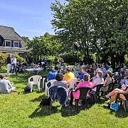 Menschen sitzen auf Stühlen im Garten bei einem Konzert mit Musikern und Sprechern