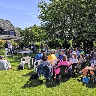 Menschen sitzen auf Stühlen im Garten bei einem Konzert mit Musikern und Sprechern